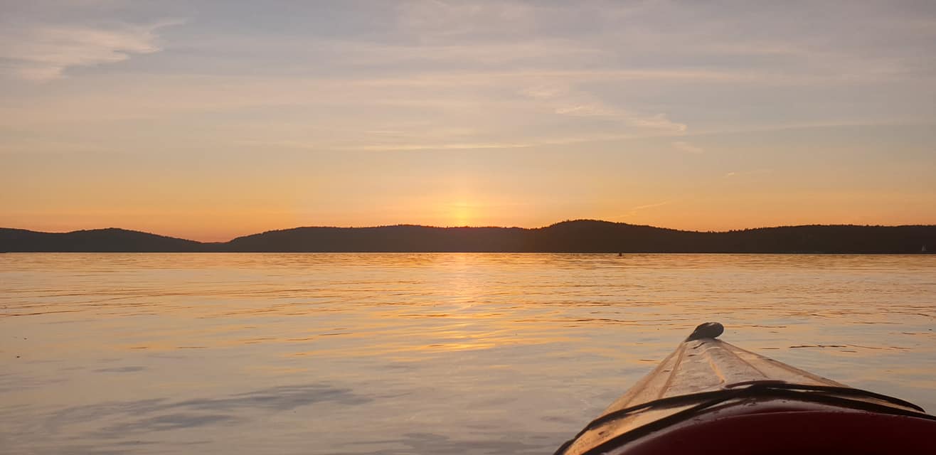 Bow of a kayak on golden water as the sun sets behind the La Cloche hills