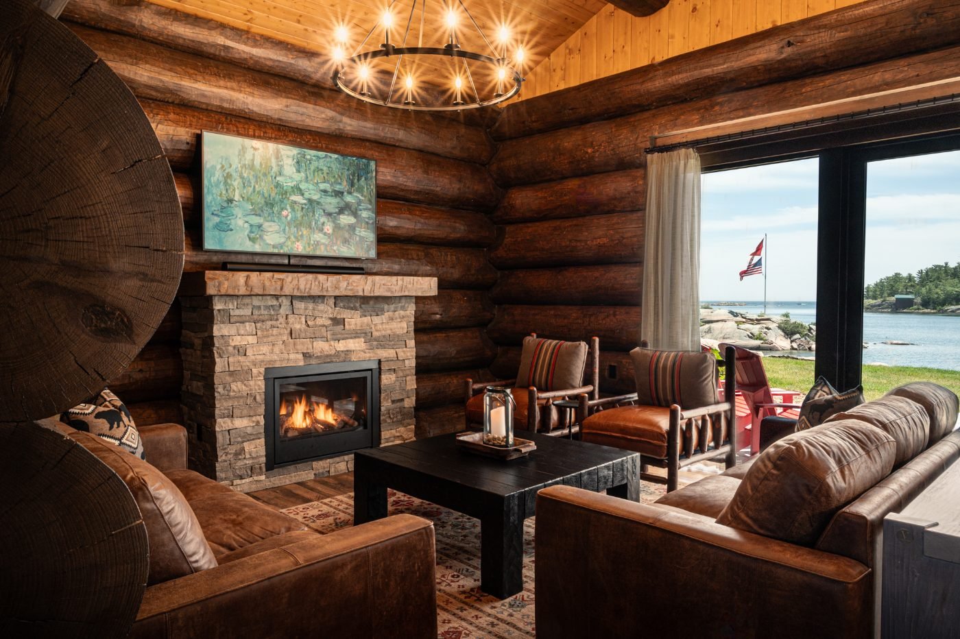 A waterfront log cabin great room with a lit stone fireplace and a glass wall onto the bay